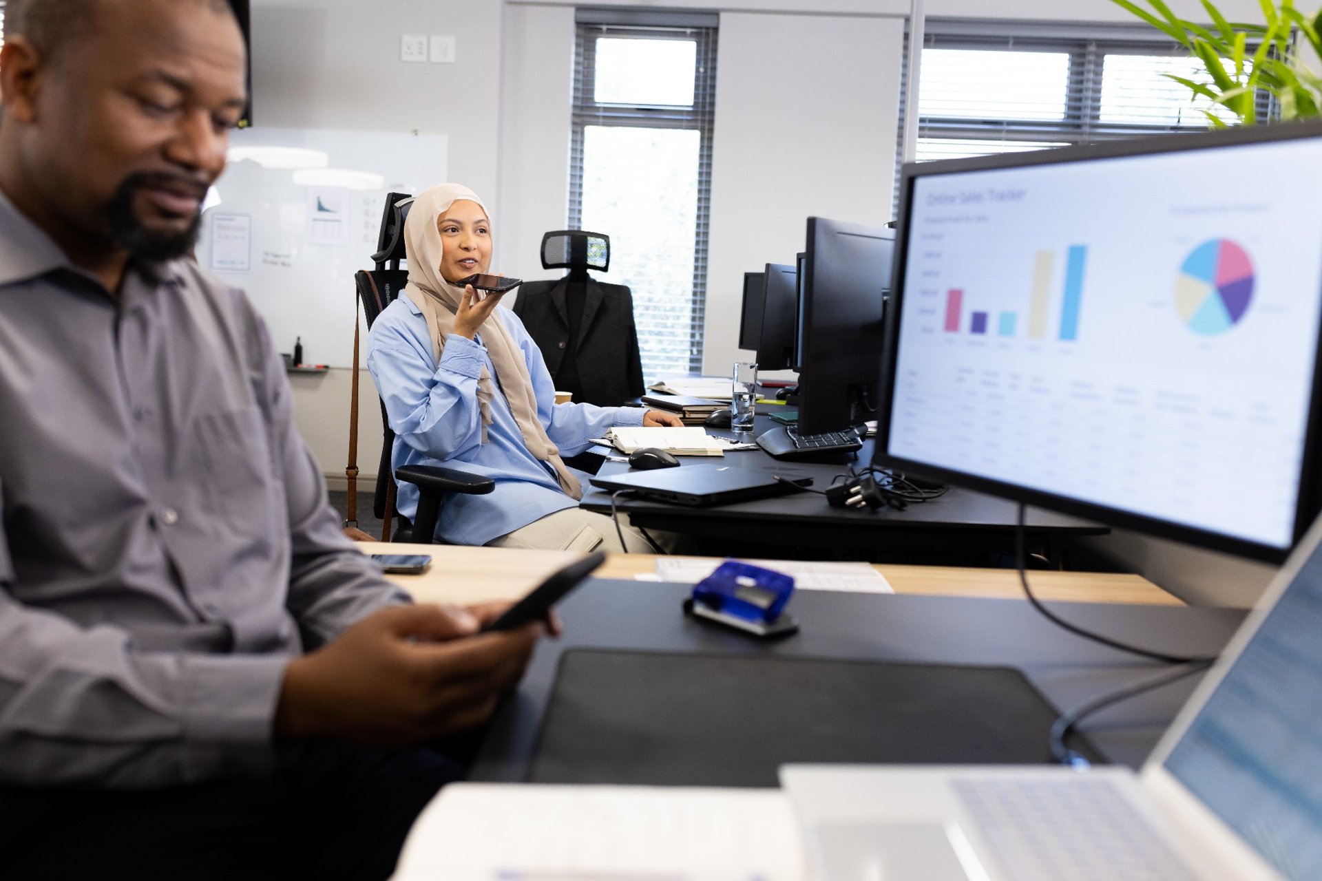 Businesswoman using voice assistant on phone at a modern office desk with charts