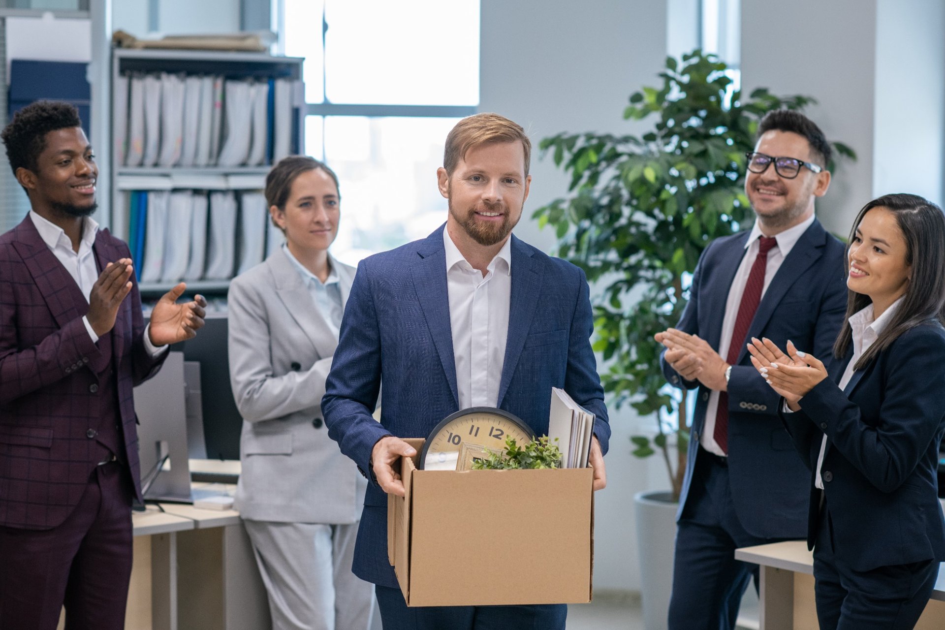 Man holding a box of personal items as coworkers clap during farewell